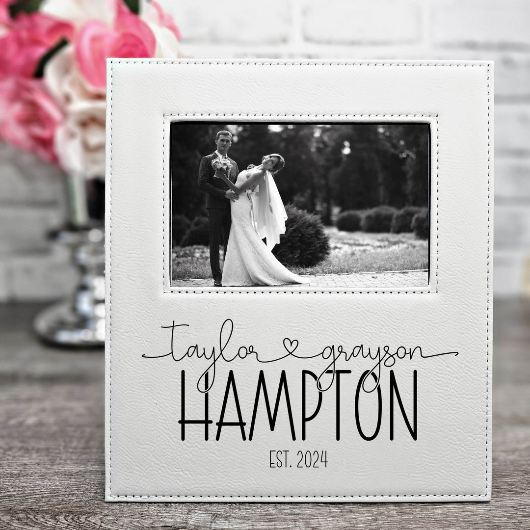 White faux leather picture frame with a black and white photo of a couple and text 'Taylor & Grayson Hampton EST. 2024' on a wooden surface. First names are in a modern script font connected with a heart. Text is engraved in black.
