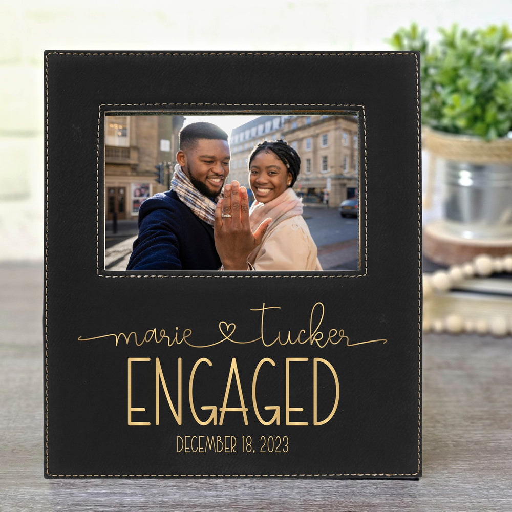 Black faux leather photo frame with 'marie & tucker ENGAGED' text along with the date engraved in metallic gold, featuring a couple on a blurred street background. Names are in modern script font and connected with a heart.