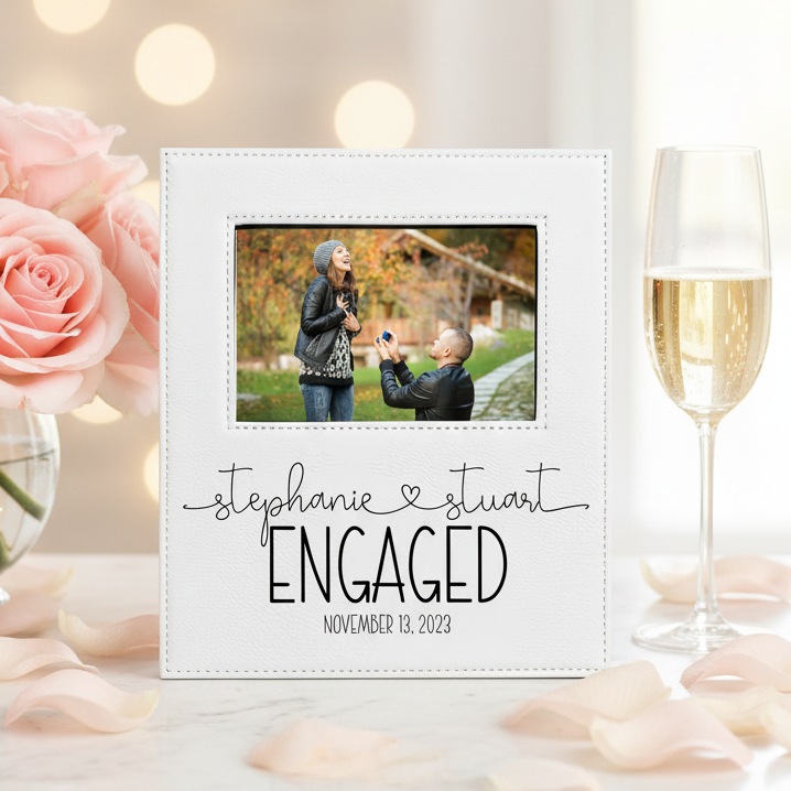 A personalized white square photo frame with a printed couple's photo and the text 'stephanie & stuart ENGAGED NOVEMBER 13, 2023,' styled on a marble surface with pink roses, scattered petals, and a champagne flute in a softly lit indoor setting.