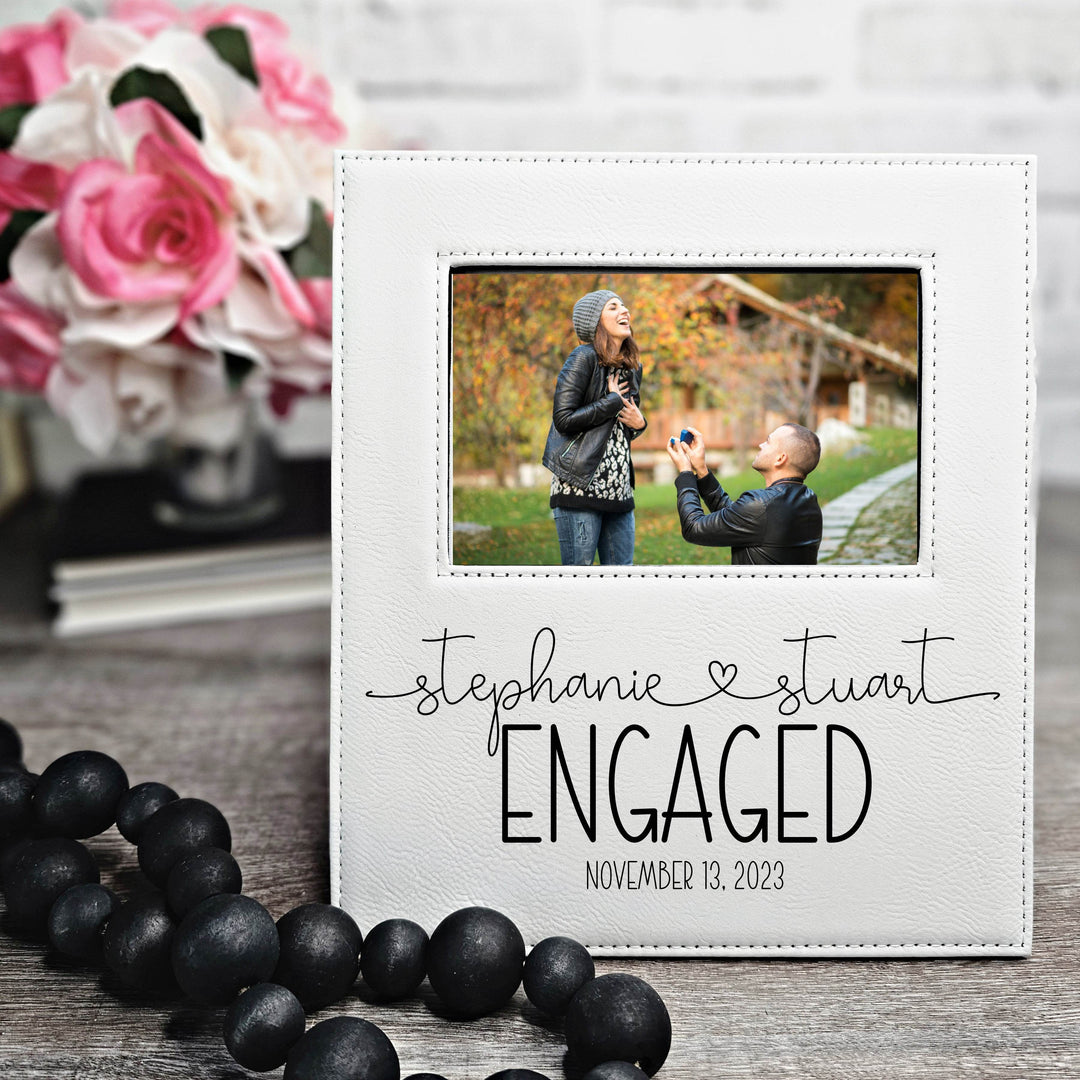 White leatherette engagement photo frame with black engraved text 'Stephanie & Stuart Engaged' along with the date. Names are connected with a heart in a modern script font.