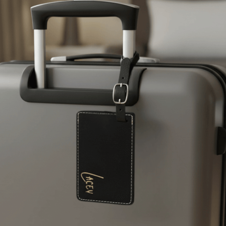 Gray suitcase with a black luggage tag featuring a gold name engraved on the bottom of the tag.