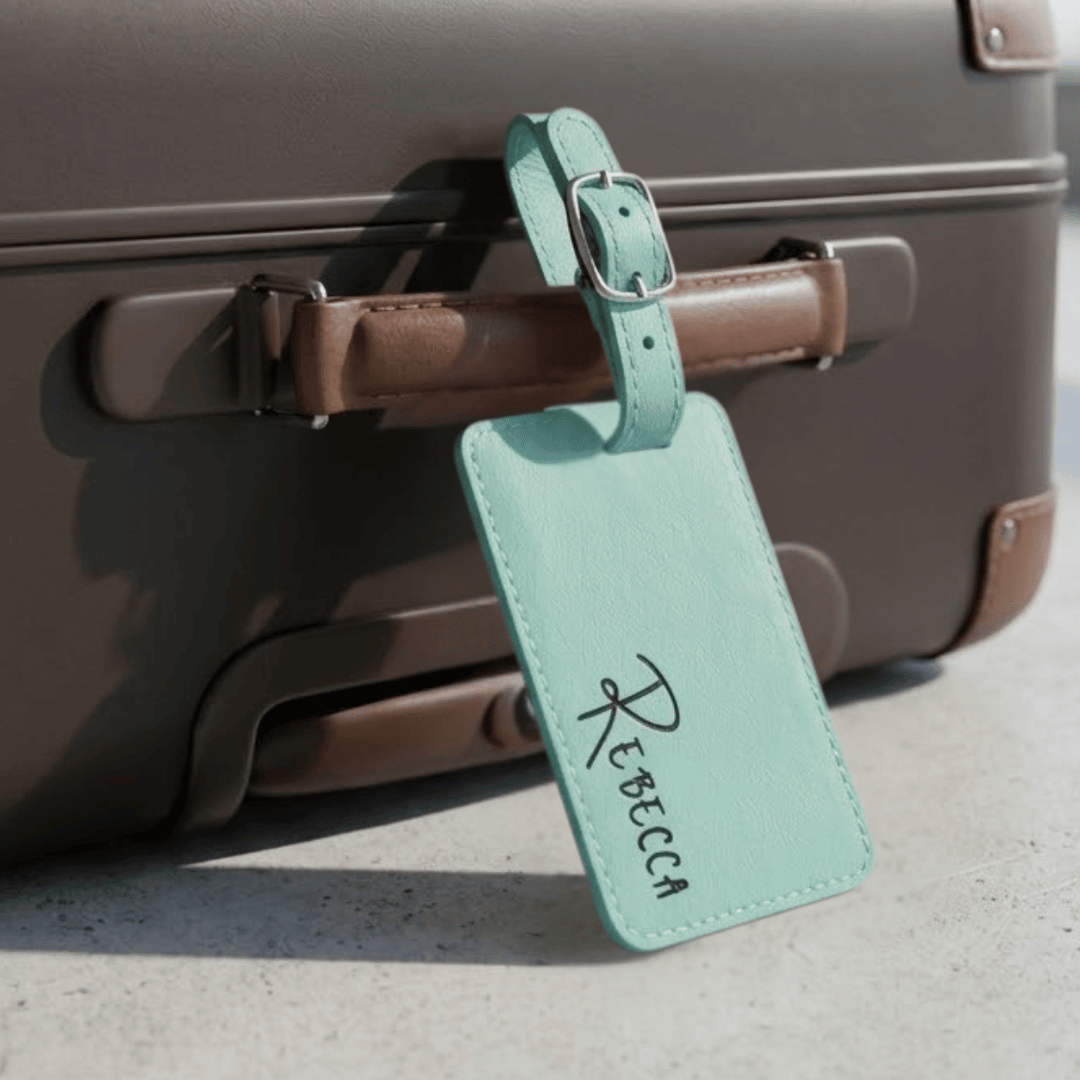 Teal luggage tag with 'Rebecca' engraved in black buckled on a suitcase.