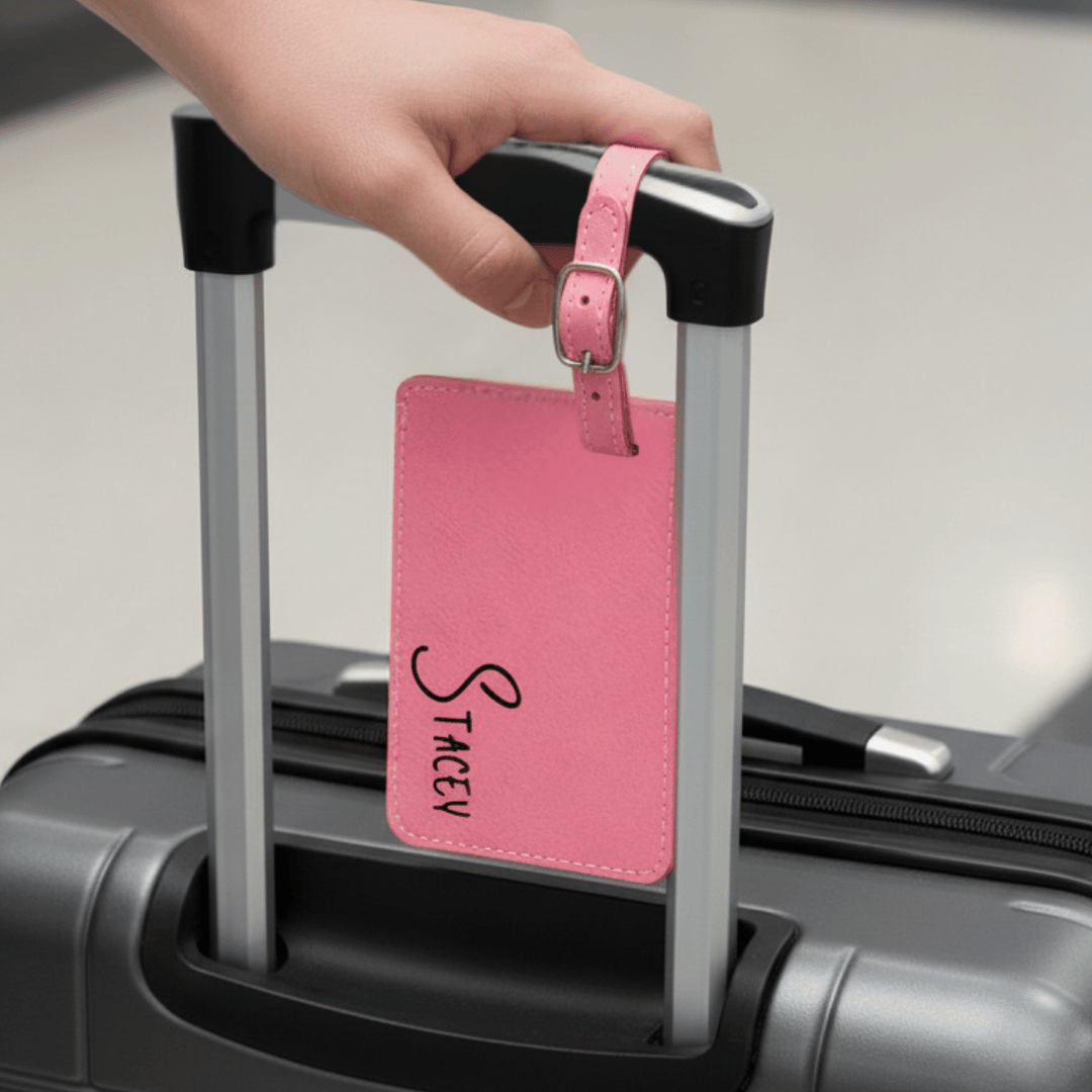 Hand holding a black suitcase with a pink luggage tag featuring the name 'Stacey' engraved on tag in an airport setting.