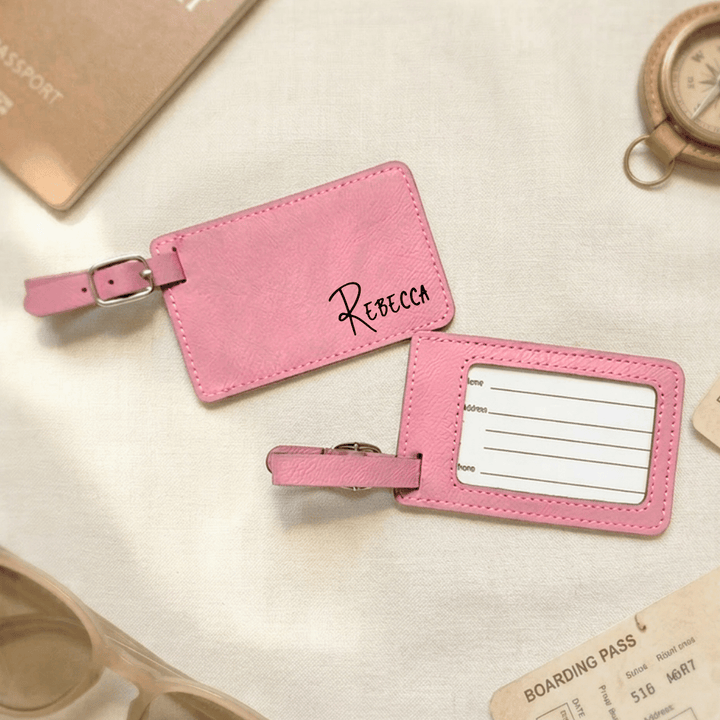 Pink luggage tags with 'Rebecca' engraved on the front in a black handwritten font and a clear plastic opening on back with place to write personal information, placed on a light surface with travel-related items.