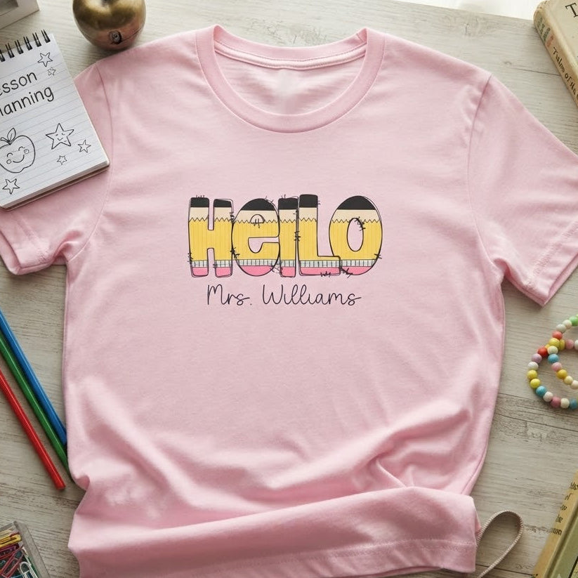 A light pink short-sleeve t-shirt with 'Hello Mrs. Williams' printed on the front in a colorful striped font, styled in a flat lay with school and classroom-themed props on a light wood background.