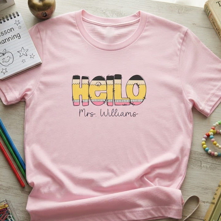 A light pink short-sleeve t-shirt with 'Hello Mrs. Williams' printed on the front in a colorful striped font, styled in a flat lay with school and classroom-themed props on a light wood background.