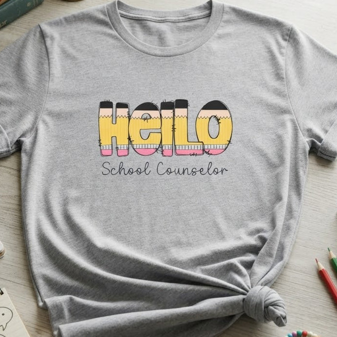 A heather gray graphic T-shirt laid flat on a light wood desk with a tied hem, featuring the words 'Hello School Counselor' in colorful pencil lettering. The shirt is styled with school-themed props including notebooks, pencils, paper clips, bracelets, and books.