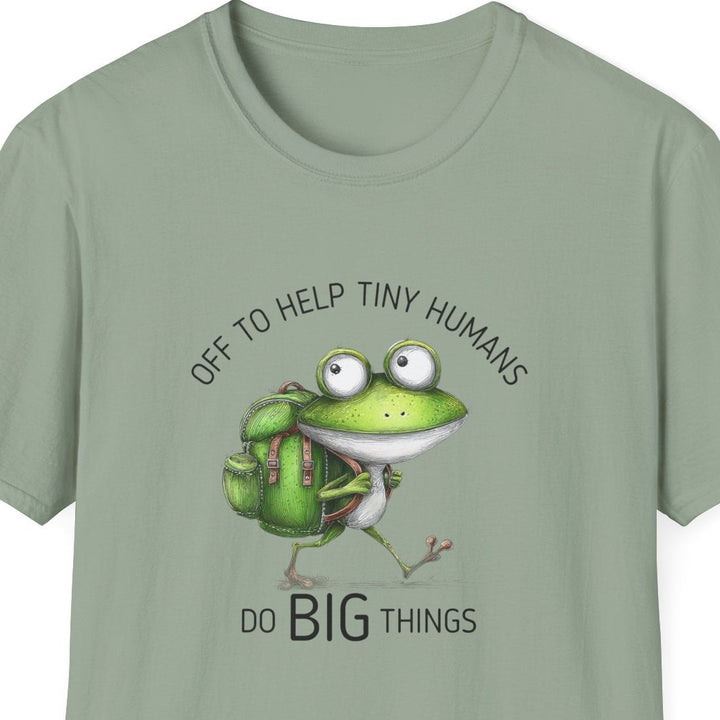 A sage green short-sleeve t-shirt with a cartoon graphic of a green frog wearing a backpack and carrying groceries, with the text 'OFF TO HELP TINY HUMANS' above and 'DO BIG THINGS' below.