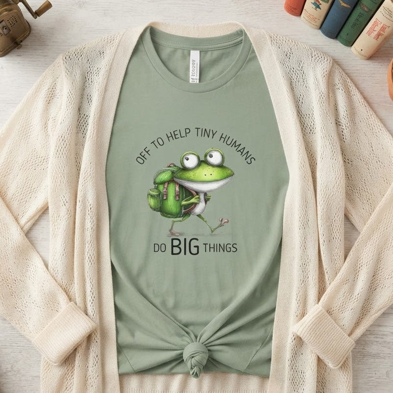 A styled flat-lay of a sage green graphic T-shirt under an open cream-colored knit cardigan. The graphic features a cartoon green frog with a backpack and the text 'OFF TO HELP TINY HUMANS DO BIG THINGS'.