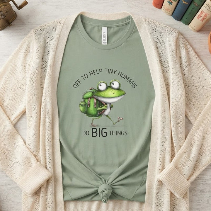 A styled flat-lay of a sage green graphic T-shirt under an open cream-colored knit cardigan. The graphic features a cartoon green frog with a backpack and the text 'OFF TO HELP TINY HUMANS DO BIG THINGS'.