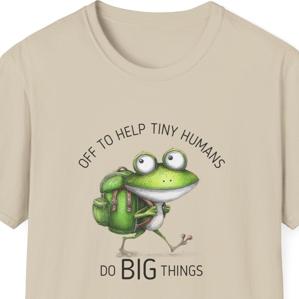A beige short-sleeve crewneck t-shirt featuring a cartoon graphic of a green frog wearing a backpack and carrying a small white bird, with the text 'OFF TO HELP TINY HUMANS' above and 'DO BIG THINGS' below.