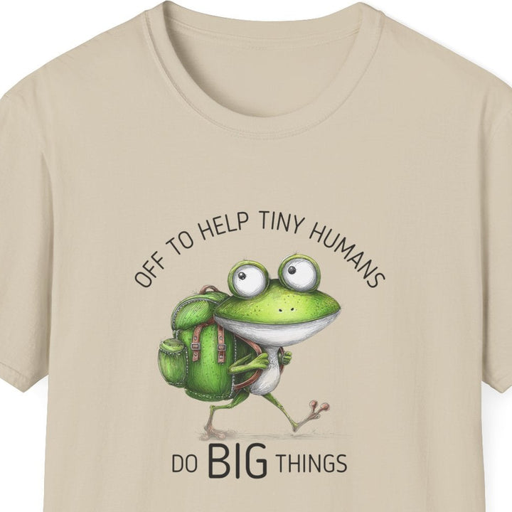 A beige short-sleeve crewneck t-shirt featuring a cartoon graphic of a green frog wearing a backpack and carrying a small white bird, with the text 'OFF TO HELP TINY HUMANS' above and 'DO BIG THINGS' below.
