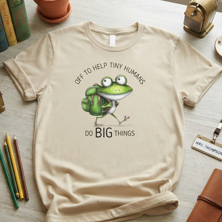 A beige graphic t-shirt featuring a cartoon green frog with a backpack and the text 'OFF TO HELP TINY HUMANS DO BIG THINGS', styled in a flat-lay on a light wooden desk surrounded by books, a succulent, a vintage camera, pencils, and a notebook.