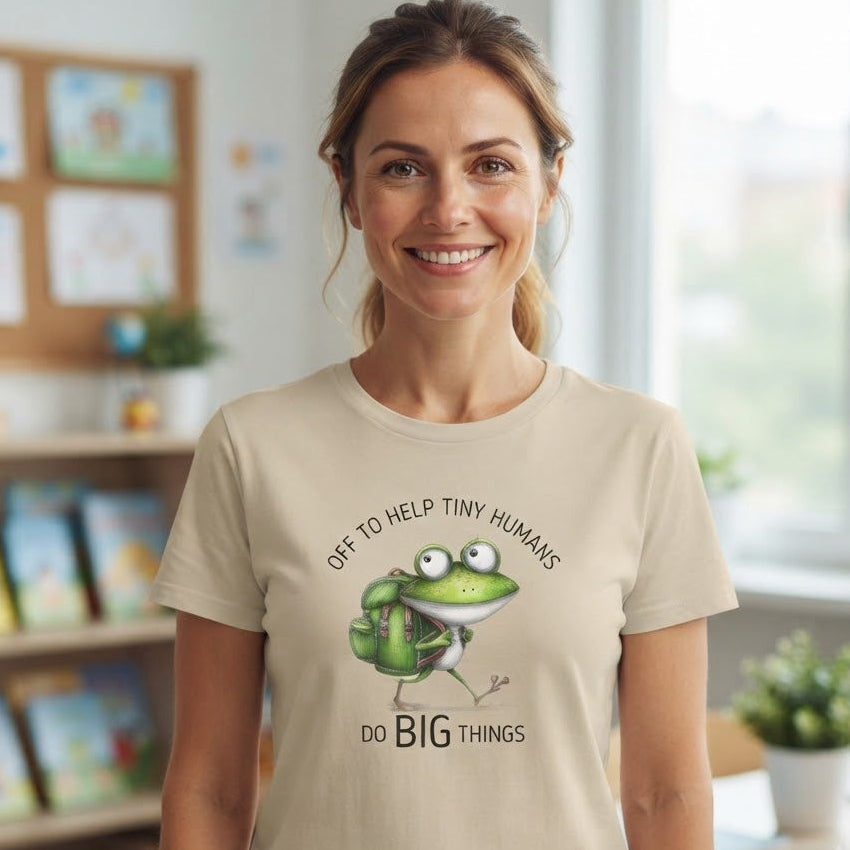 A smiling woman stands in a bright classroom wearing a light beige graphic T-shirt. The shirt features a cartoon green frog with a backpack carrying books, with the text 'OFF TO HELP TINY HUMANS' above and 'DO BIG THINGS' below.
