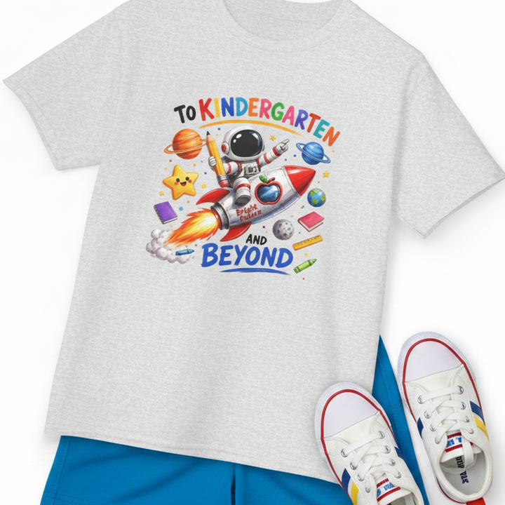 A kids' heather ash gray short-sleeve T-shirt with a colorful space-themed 'To Kindergarten and Beyond' graphic, styled flat-lay with blue shorts and white sneakers on a white background.