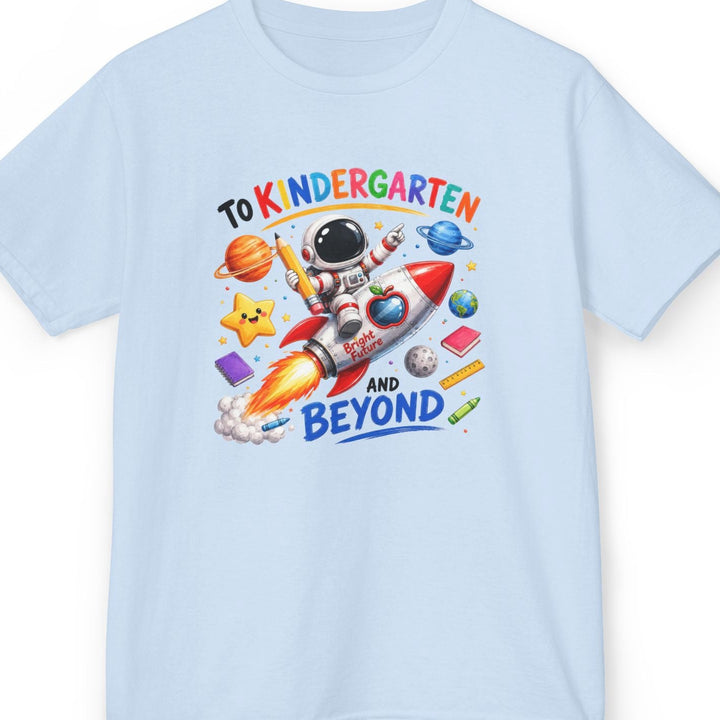 A light blue short-sleeve t-shirt with a colorful cartoon graphic of an astronaut riding a rocket ship, surrounded by planets, stars, and school supplies, with the text 'TO KINDERGARTEN AND BEYOND'.
