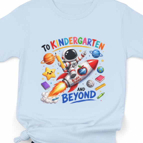 A light blue short-sleeve t-shirt with a colorful cartoon graphic on the front. The graphic features an astronaut riding a rocket ship with the text 'TO KINDERGARTEN AND BEYOND' and various school and space-themed icons like planets, stars, and books.