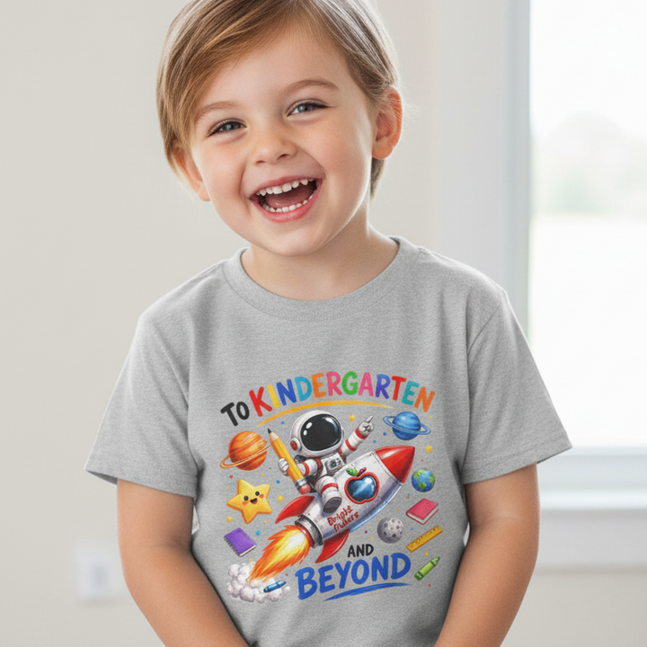 A smiling young boy wearing a light gray t-shirt with a colorful space-themed graphic that reads 'TO KINDERGARTEN AND BEYOND' with an astronaut, rocket, planets, stars, and school icons.
