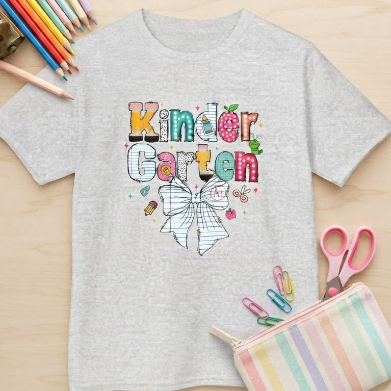 A light ash gray short-sleeve t-shirt with a colorful 'Kindergarten' text graphic and a bow illustration on the front, styled in a flat-lay on a light wood background with school supplies including colored pencils, a pencil sharpener, scissors, and a pastel striped pencil pouch.