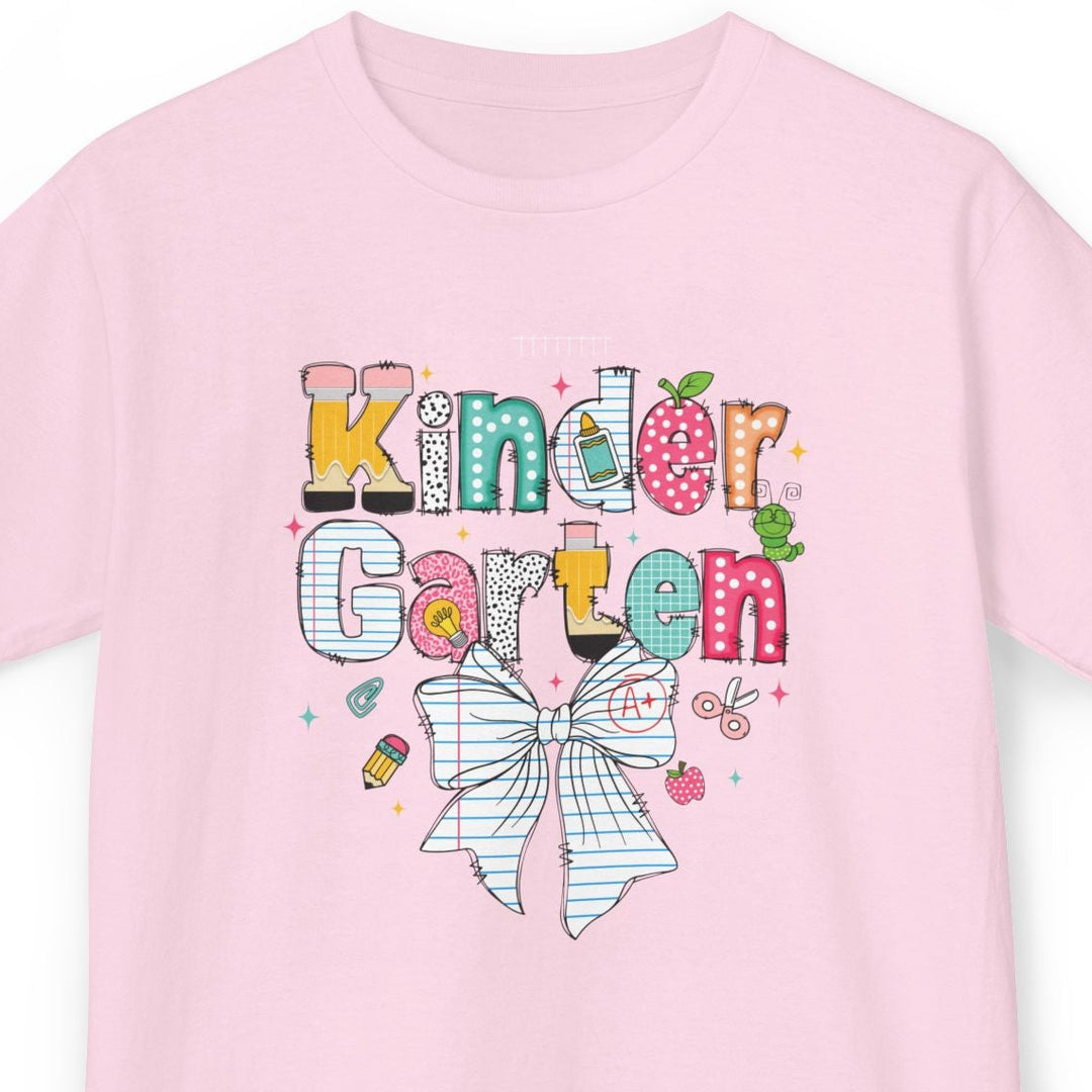 A light pink short-sleeve t-shirt with a colorful graphic on the front that reads 'Kinder Garten' in playful multicolored letters above a large white bow illustration, shown on a plain white background.