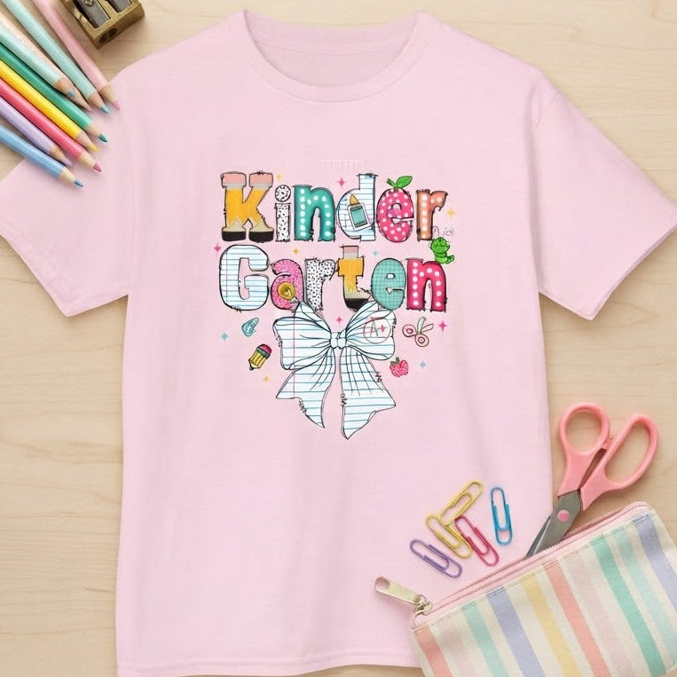 A light pink short-sleeve t-shirt with a colorful 'Kindergarten Garten' graphic on the chest, styled in a flat-lay on a light wood background with school supplies including colored pencils, a pencil sharpener, scissors, paper clips, and a striped pencil pouch.