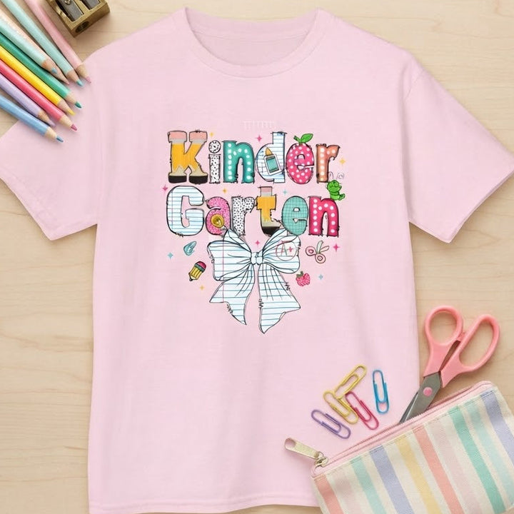 A light pink short-sleeve t-shirt with a colorful 'Kindergarten Garten' graphic on the chest, styled in a flat-lay on a light wood background with school supplies including colored pencils, a pencil sharpener, scissors, paper clips, and a striped pencil pouch.