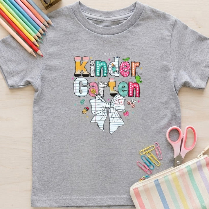 A heather gray short-sleeve kids' T-shirt laid flat on a light wood surface, printed with the words 'Kinder Garten' in colorful playful letters and a white bow graphic. Surrounding props include colored pencils, a pencil sharpener, pink-handled scissors, and a pastel striped pencil pouch.
