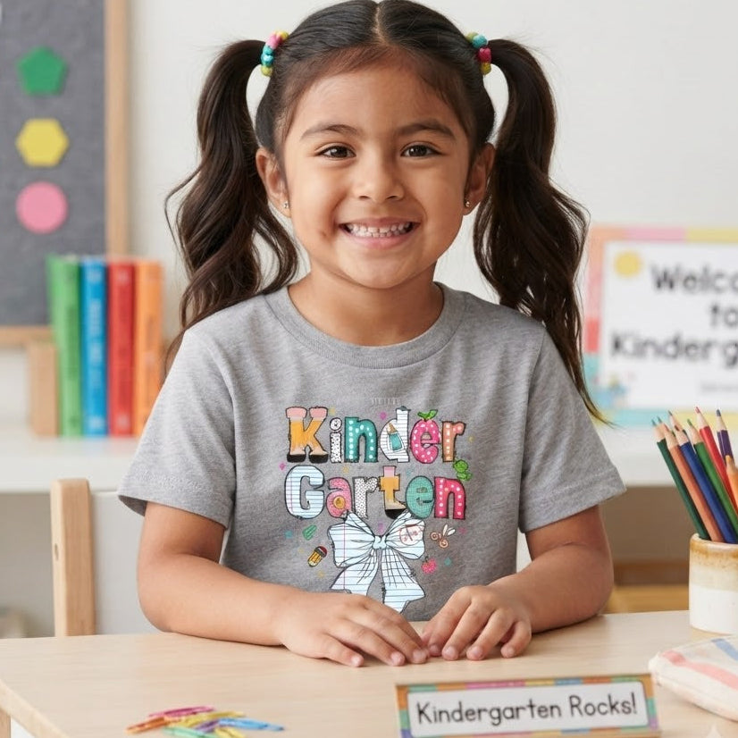 A young girl with dark hair in pigtails sits at a wooden desk in a bright classroom, wearing a gray t-shirt with a colorful 'Kinder Garten' graphic and a white bow illustration. The desk has a nameplate that reads 'Kindergarten Rocks!' and is surrounded by school supplies.