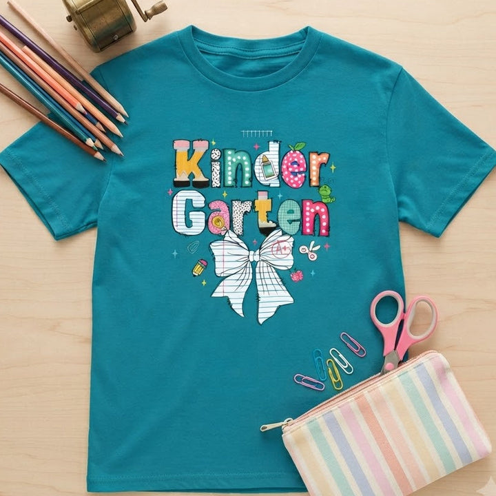 A teal short-sleeve kids' T-shirt laid flat on a light wood surface, printed with colorful 'Kinder Garten' lettering and a white bow graphic. Surrounding props include colored pencils, a brass pencil sharpener, pink scissors, paper clips, and a pastel striped zip pouch.