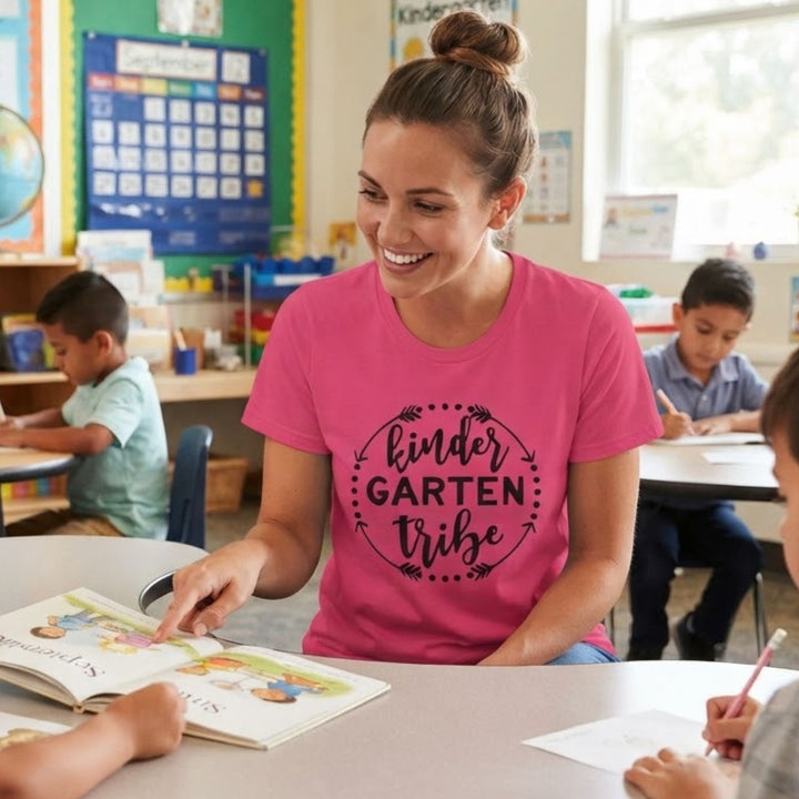 A smiling woman in a pink graphic t-shirt sits at a desk in a classroom, pointing to a children's book. The shirt features the text 'kinder GARTEN tribe' in black.