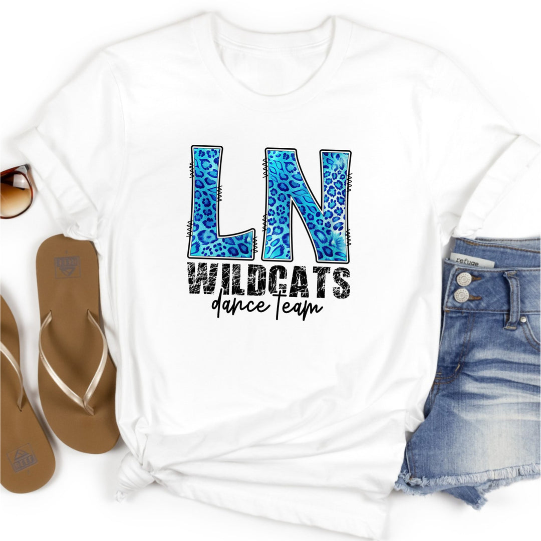 White t-shirt with 'LN Wildcats dance Team' design, surrounded by sandals and denim shorts on a white background. The 'LN' is in large blue leopard doodle letters and 'wildcats' is written in a cat scratched font. 'Dance team' is underneath in a modern script font.