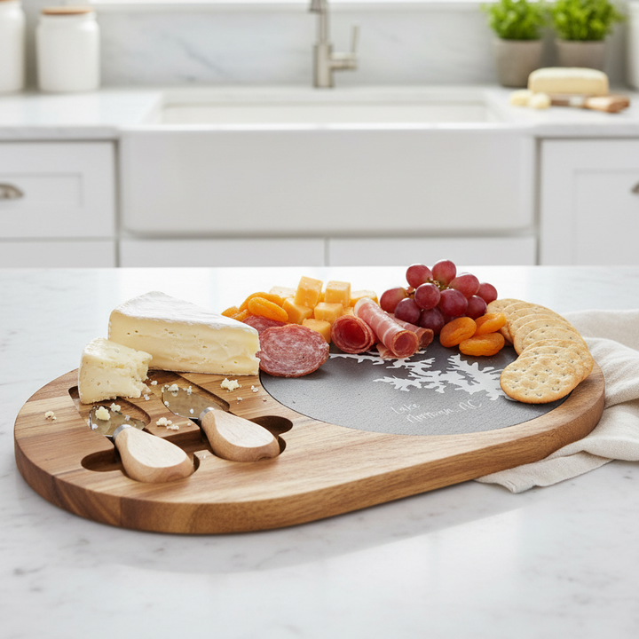 Oval wooden charcuterie board with cheese, fruits, and meats on a kitchen counter. It's made of acacia wood and slate. The slate has silhouette of Lake Norman, NC engraved on it.