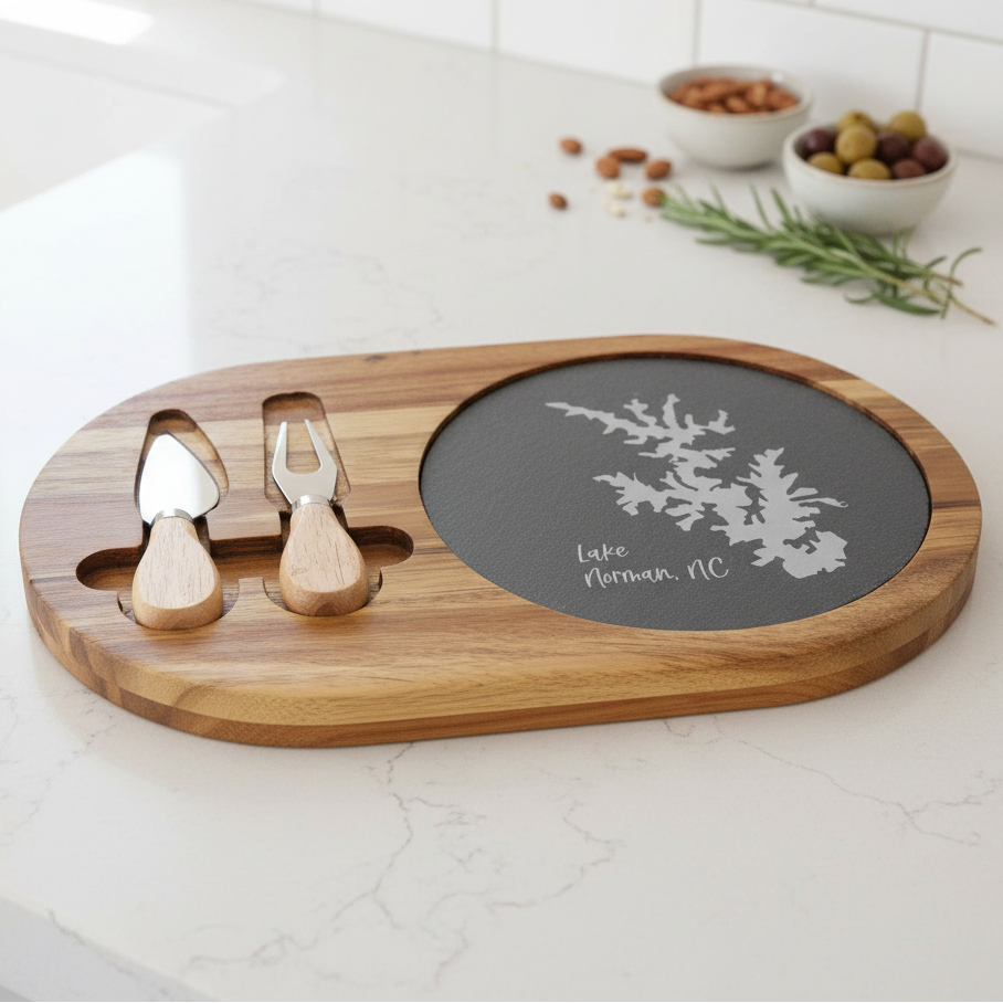 Wooden cheese board with slate center and 2 serving utensils on a kitchen counter. Slate has silhouette of Lake Norman, NC along with the words engraved in slate