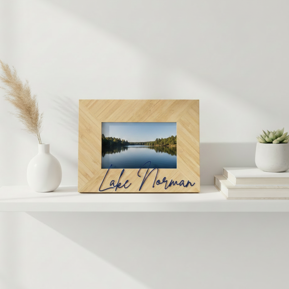 A personalized wooden bamboo picture frame with a herringbone pattern, featuring a landscape photo of a lake and the text 'Lake Norman' in navy blue script. The frame is styled on a white shelf with decorative props including a vase with dried grass, books, and a small potted succulent.