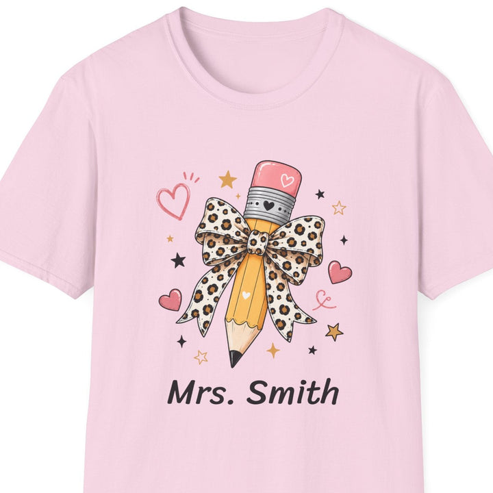 A light pink short-sleeve crewneck t-shirt featuring a graphic of a pencil with a leopard-print bow, surrounded by small hearts and stars, with the text 'Mrs. Smith' printed below.