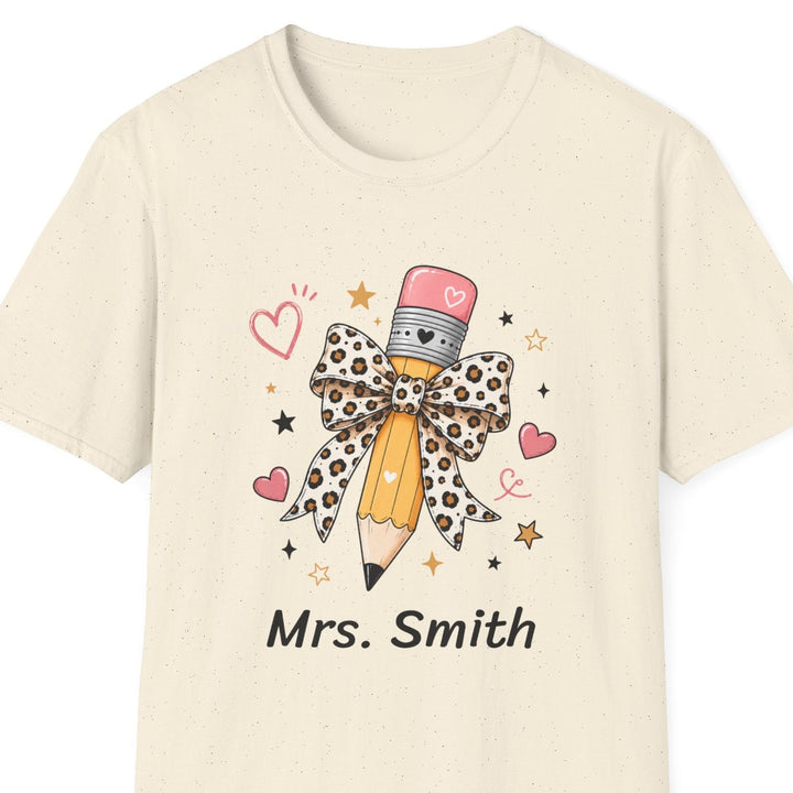 A cream-colored short-sleeve t-shirt with a graphic of a pencil decorated with a leopard-print bow, surrounded by pink hearts and gold stars, with the text 'Mrs. Smith' printed below.