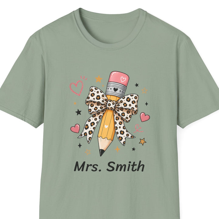 A sage green short-sleeve t-shirt with a custom graphic of a pencil wrapped in a leopard-print bow, surrounded by pink hearts and stars, with the text 'Mrs. Smith' printed below.