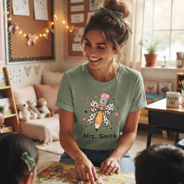 A woman in a classroom wearing a personalized sage green graphic T-shirt with a pencil illustration and the text 'Mrs. Smith'.