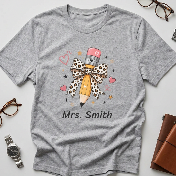 A heather gray short-sleeve T-shirt laid flat on a light gray background, styled with eyeglasses, a watch, a brown leather notebook, and a small bottle around it. The shirt has a graphic of a pencil with a leopard-print bow, pink hearts, stars, and the text 'Mrs. Smith'