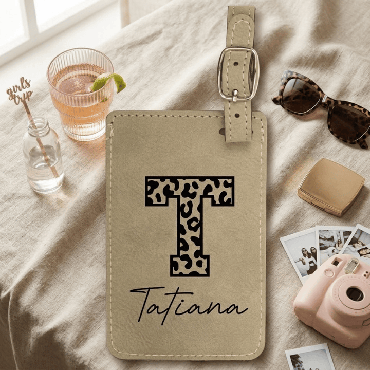 Beige luggage tag with leopard print letter 'T' and name 'Tatiana' engraved in black on a light fabric background with a drink, sunglasses, and camera.