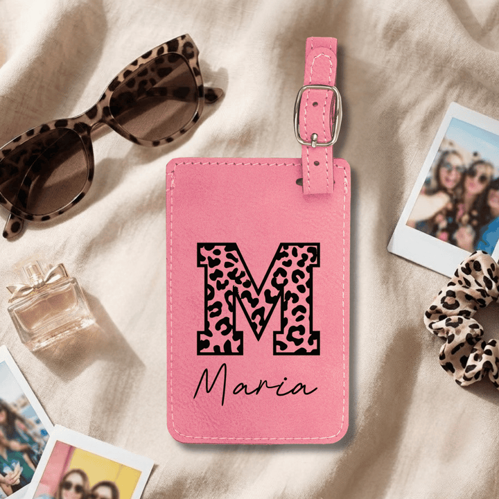 Pink luggage tag with leopard print letter 'M' and name 'Maria' engraved in black, surrounded by sunglasses, perfume, and photos on a beige surface.