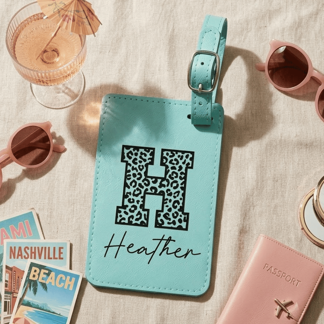 Turquoise luggage tag with leopard print letter 'H' and name 'Heather' engraved in black, surrounded by travel items on a beige surface.