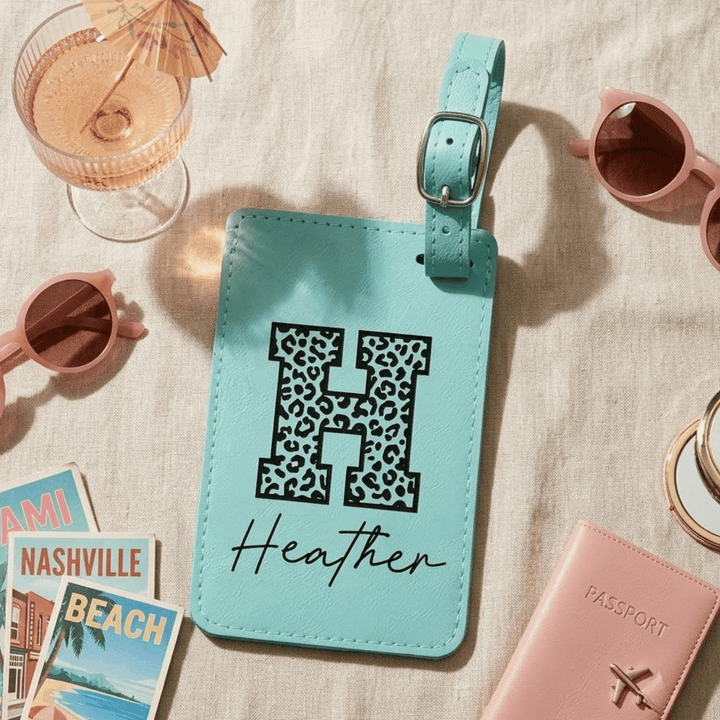 Turquoise luggage tag with leopard print letter 'H' and name 'Heather' engraved in black, surrounded by travel items on a beige surface.