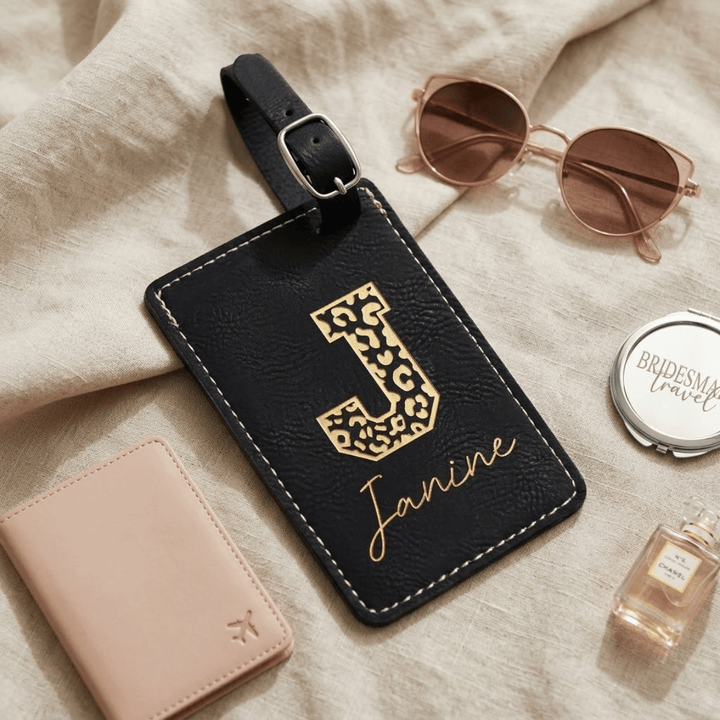 Black leather luggage tag with gold letter 'J' in leopard print and name 'Janine', sunglasses, wallet, and perfume bottle on a beige fabric background.