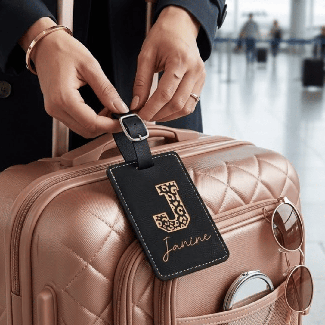 Person attaching a luggage tag with 'Janine' and leopard 'J' engraved in metallic gold on a pink suitcase in an airport setting
