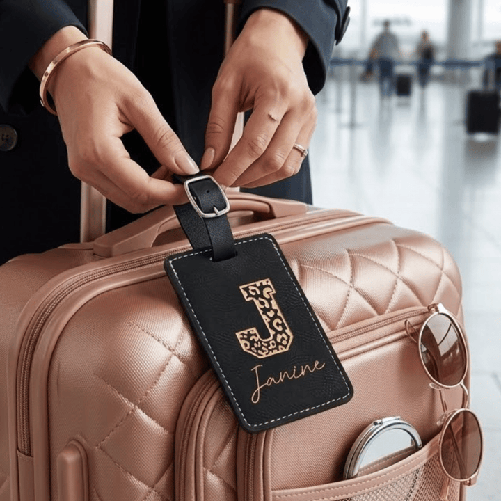 Person attaching a luggage tag with 'Janine' and leopard 'J' engraved in metallic gold on a pink suitcase in an airport setting