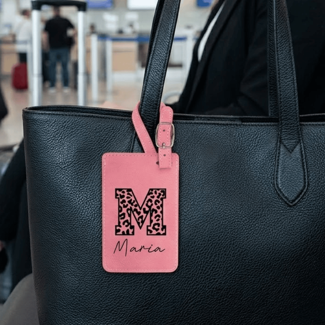 Black handbag with a pink luggage tag featuring a leopard print letter 'M' and name 'Maria' engraved in black.
