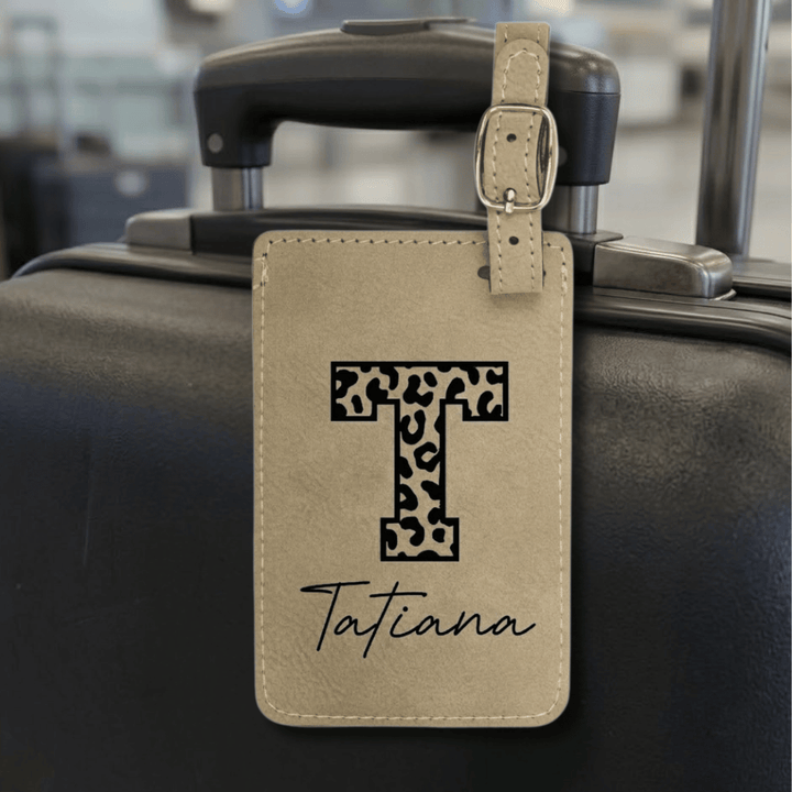 Luggage tag with leopard print letter 'T' and name 'Tatiana' engraved in black attached to a suitcase.