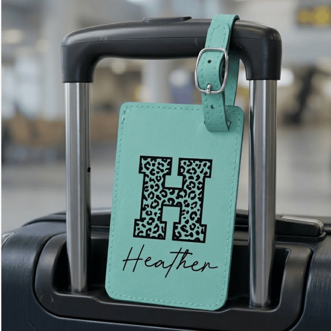 Teal luggage tag with 'H' in leopard print and 'Heather' engraved in black on the tag attached on a suitcase handle.