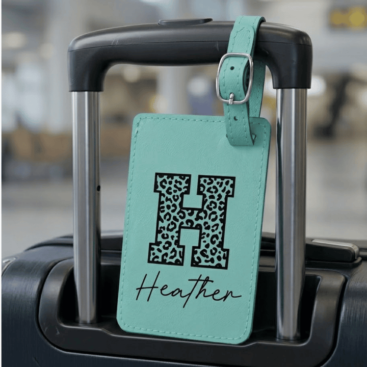Teal luggage tag with 'H' in leopard print and 'Heather' engraved in black on the tag attached on a suitcase handle.